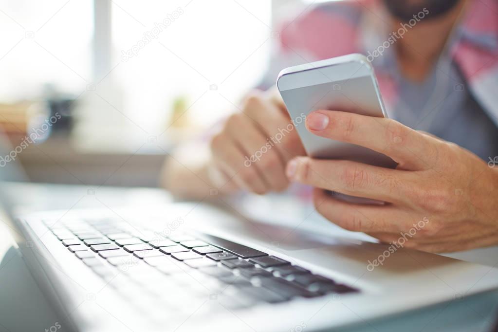 Close-up of unrecognizable man texting message on smartphone when he working at desk with laptop