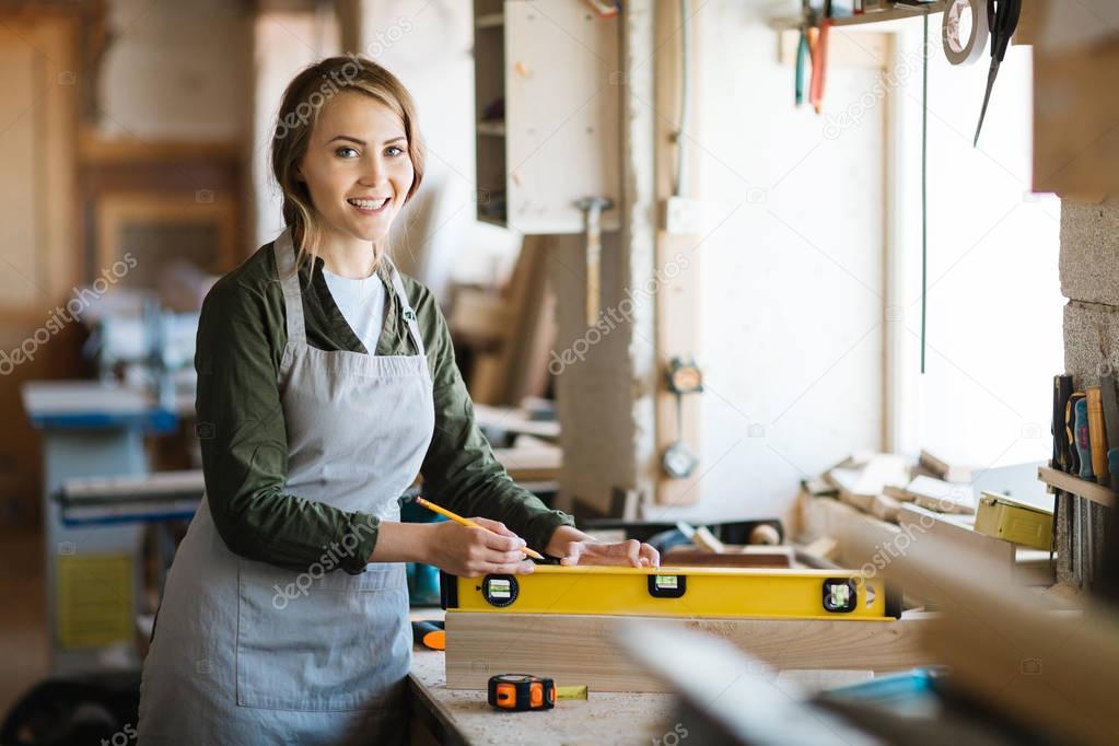 Portrait of pretty young carpenter in apron looking at camera with wide smile while holding spirit level on plank, blurred background