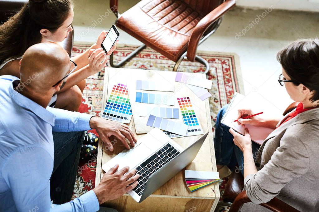 High angle view of busy interior designers brainstorming on promising project while sitting around wooden table covered with color swatches