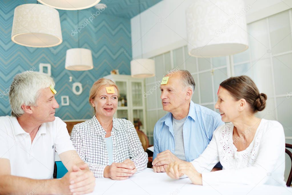 Cheerful elderly friends playing guessing game while sitting at table with sticky notes on their foreheads, interior of stylish cafe on background