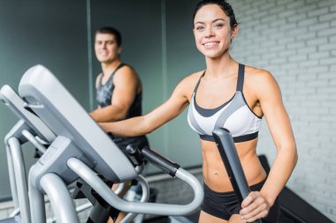 Eliptik sonraki adam, hem de gülümseyerek kameraya modern spor olarak çalışma sırasında uyacak makinasıyla egzersiz güzel sportif esmer kadın portresi
