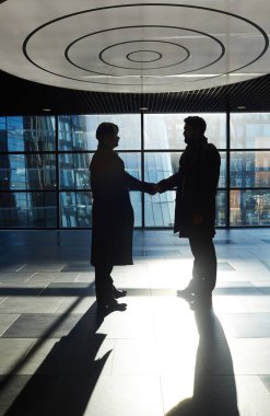 Siluetleri panoramik pencerelere, profil görünümü ile geniş office lobide duran el sıkışan iki iş ortakları