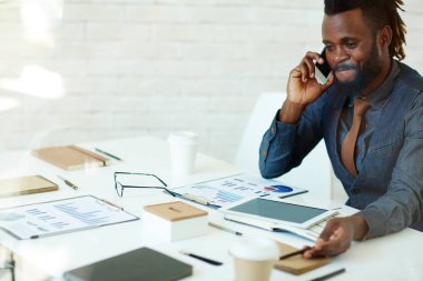 Waist-up portrait of cheerful Afro-American manager talking to his business partner on smartphone while sitting at meeting table in boardroom