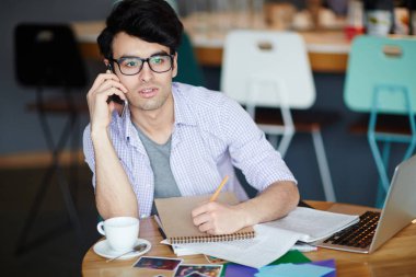 Rahat kıyafetler ve gözlük telefon ederken ve kafede dizüstü bilgisayar ile çalışma sırasında notlar yapma masada oturan genç yaratıcı adam portresi