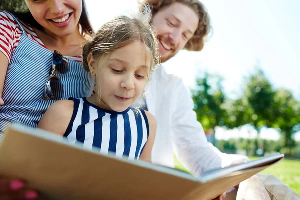 愛らしい少女と彼女の両親は 公園で一緒に面白い本を読んで — ストック写真