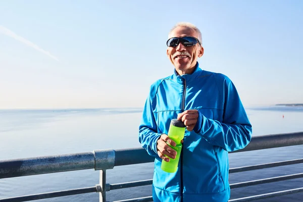 ボトル 雲一つない青空と背景に深い湖から水を飲むために屋外でのトレーニングからの短い休憩を取っている間カメラを見て笑顔のシニア スポーツマンのウエスト アップ肖像画 — ストック写真