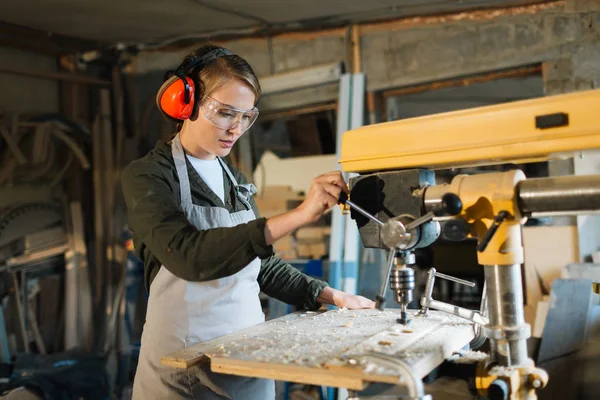 Pretty young carpenter wearing ear and eye protectors while using drill press to make holes in wooden work piece, waist-up portrait