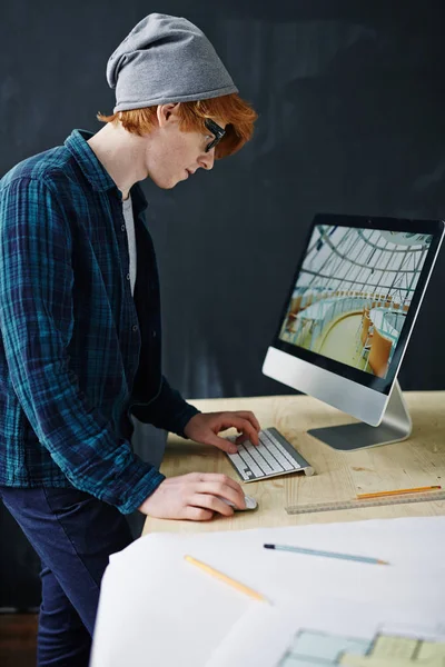 ビーニー帽子とオフィスで黒板背景に現代のコンピューターで作業しながら机に立ってメガネを身に着けている若い創造的な赤髪の男の側ビュー肖像画 — ストック写真
