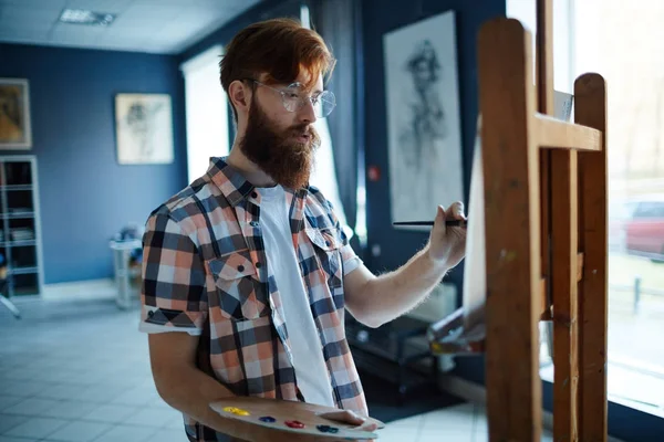 Young man with paintbrush and palette standing in front of easel in art-studio