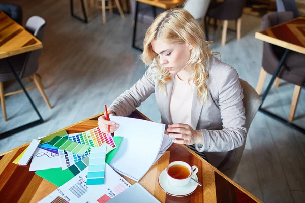 Attractive blond-haired designer working on promising project while sitting at modern cafe, cup of herbal tea and color swatches on wooden table, waist-up portrait shot