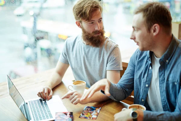 Profile view of handsome man chatting animatedly with his bearded friend while sitting in cozy coffeehouse with panoramic windows