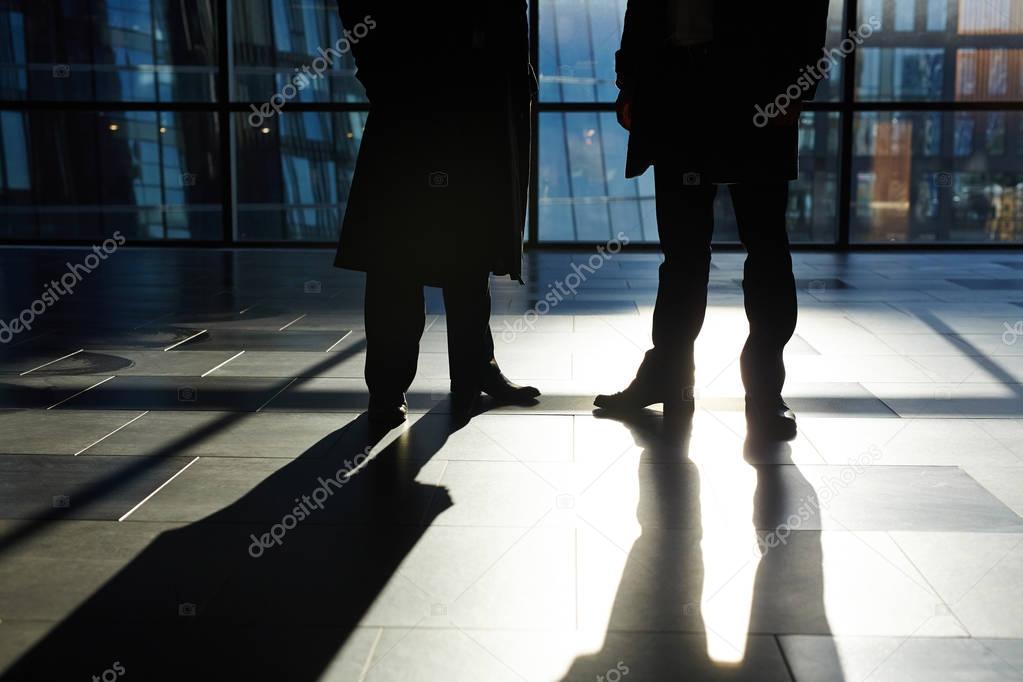 Two unrecognizable white collar workers standing against panoramic window in modern office lobby, their shadows reflecting on tile flooring