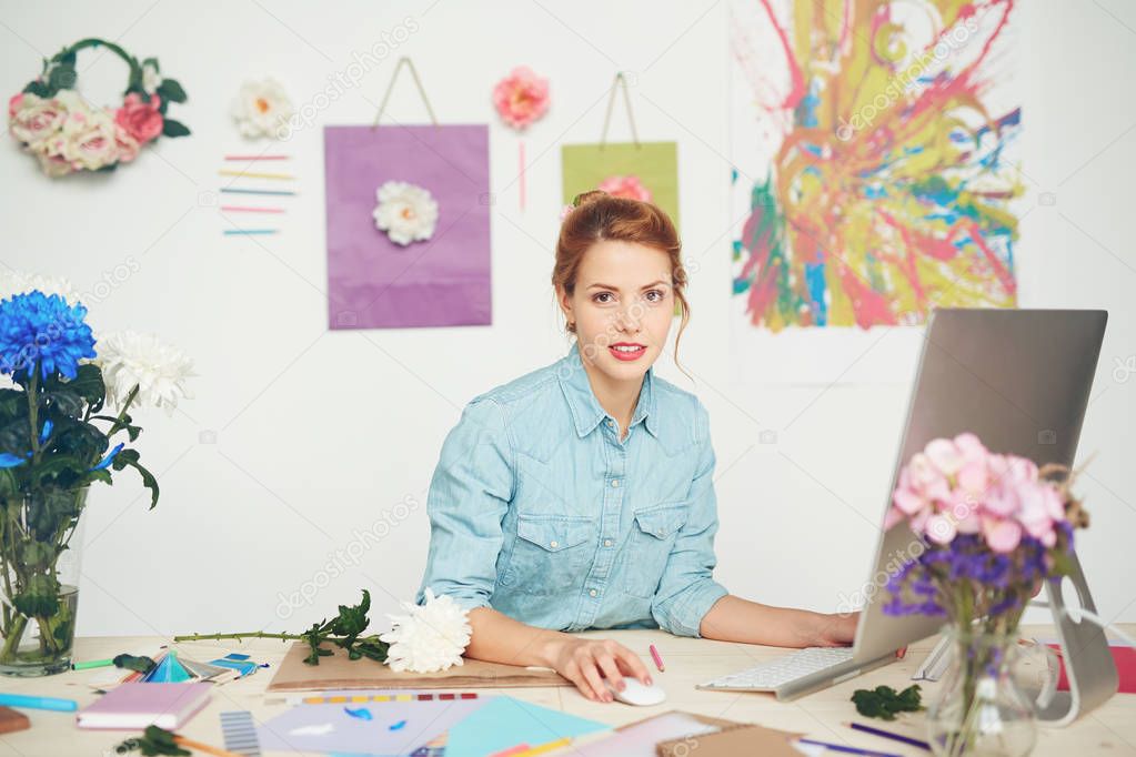 Confident young florist in denim shirt sitting in front of computer and checking emails, bouquet of blue and white chrysanthemums standing on messy table