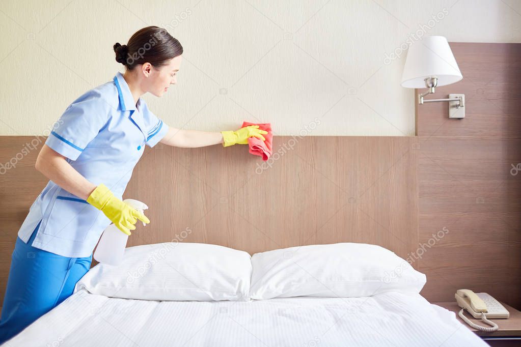 Pretty chambermaid dusting bed with sponge in hotel-room