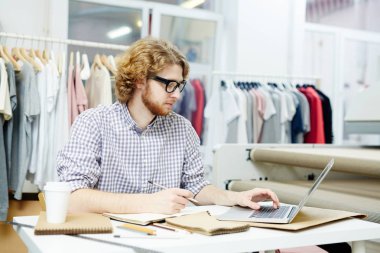 Studio dizüstü bilgisayar önünde oturan ve yapım genç çağdaş tasarımcı not defterinde skeçler