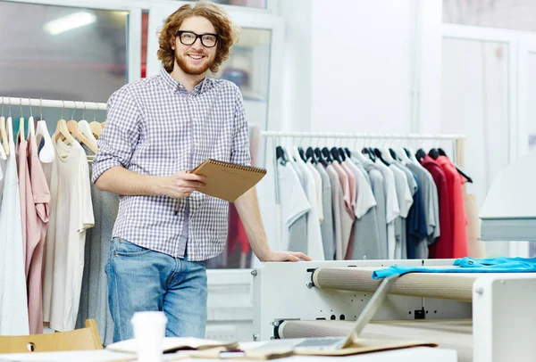 Young specialist of typography design with notepad standing by printing machine in studio