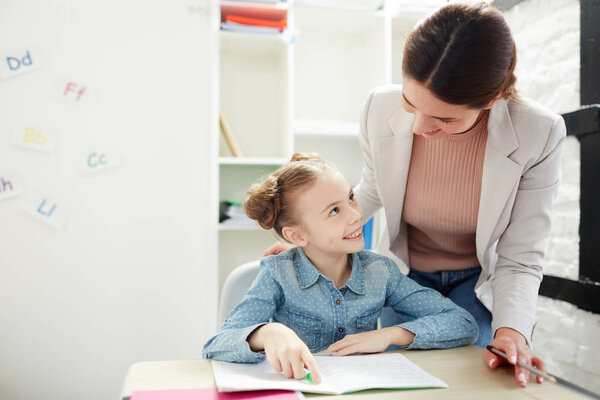 Cute little schoolgirl looking at teacher with smile while discussing notes in her copybook