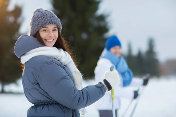 灰色のビーニー 白いスカーフと暖かい冬のジャケットを公園や森で彼氏と一緒にスキーをしながらカメラ目線で微笑んでいる女の子 — ストック写真