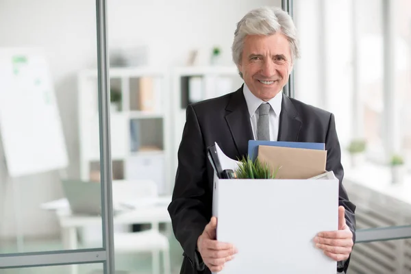 Happy senior man with box standing by his new office and looking at camera