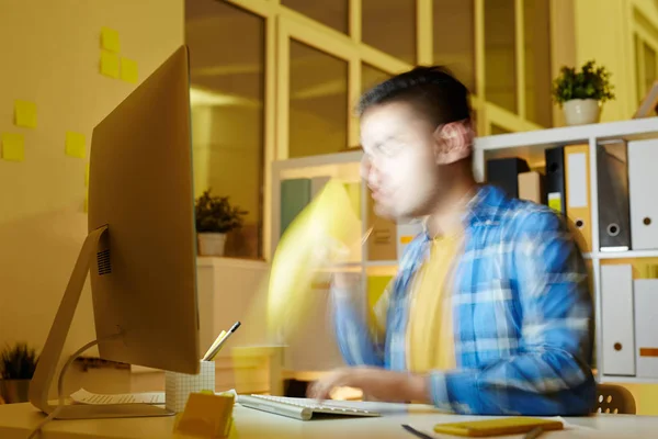 Blurry businessman drinking coffee by workplace while working in front of computer at night