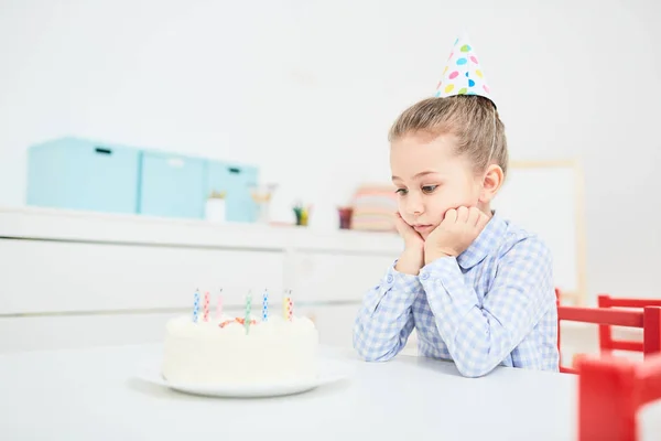 Sad little girl looking at birthday cake with candles while sitting alone by table