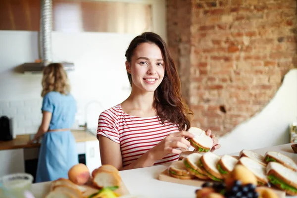 カメラ目線に立っている間提供テーブルで彼女の友人と背景にサンドイッチと幸せな女 — ストック写真