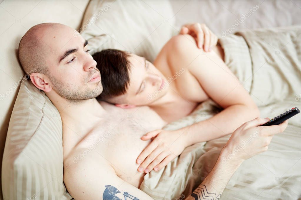 Gay couple lying in bed and watching television broadcast in the evening