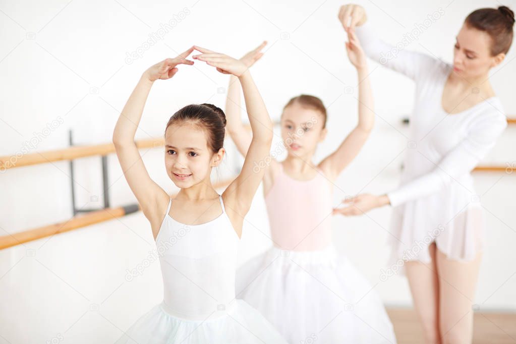 Little ballerina dancing with her arms over head while teacher helping her friend near by