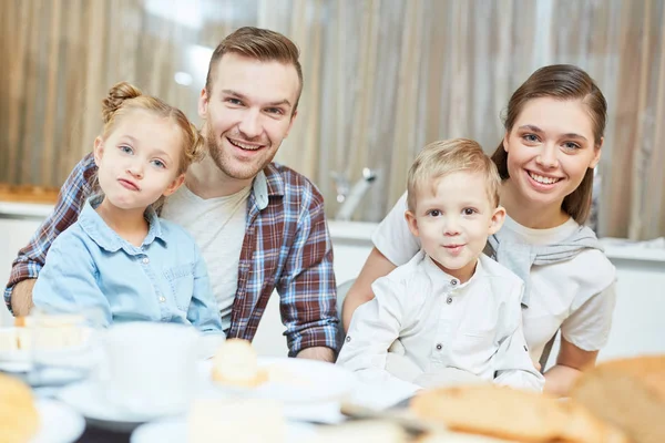 Funny siblings and their young parents looking at camera while sitting by table