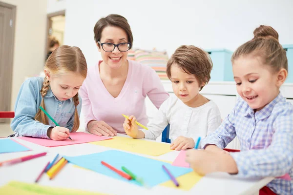 愉快的老师和三个孩子坐在桌在图画课在幼稚园 — 图库照片