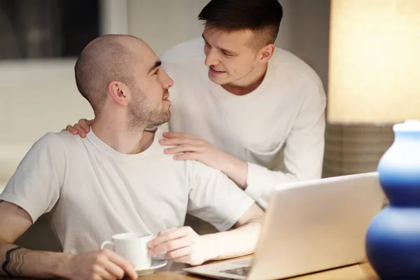 多情的年轻同性恋男子与杯饮料和笔记本电脑看着他的情人拥抱他 — 图库照片