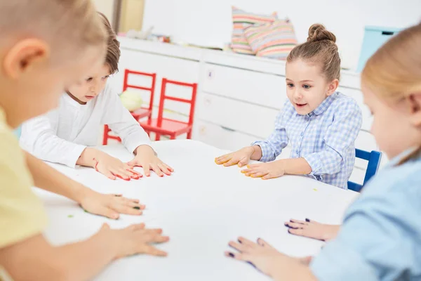 テーブルのそばに座って紙のシートに手形を作るかわいい子供たちのグループ — ストック写真