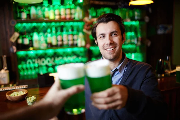 Young smiling man with beer toasting with his friend in pub during celebration of Saint Patrick day