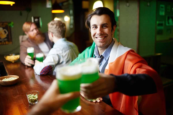 微笑的年轻人喝啤酒在他的朋友在酒吧的背景下烘烤 — 图库照片