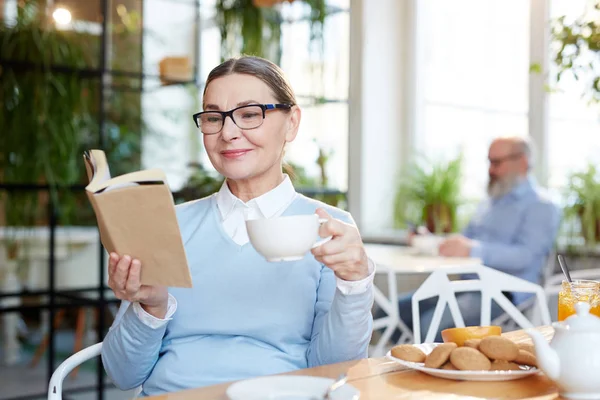 カフェでクッキーとお茶を飲んで 本を読んで現代のシニア女性 — ストック写真