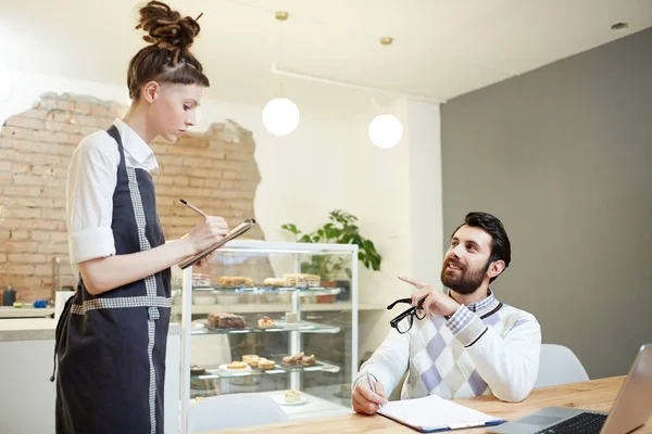 Young waitress writing down order of businessman in cafe or restaurant during lunch break