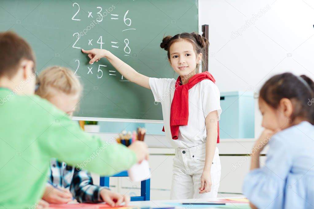 Clever girl standing by blackboard and pointing at one of sums during lesson of maths