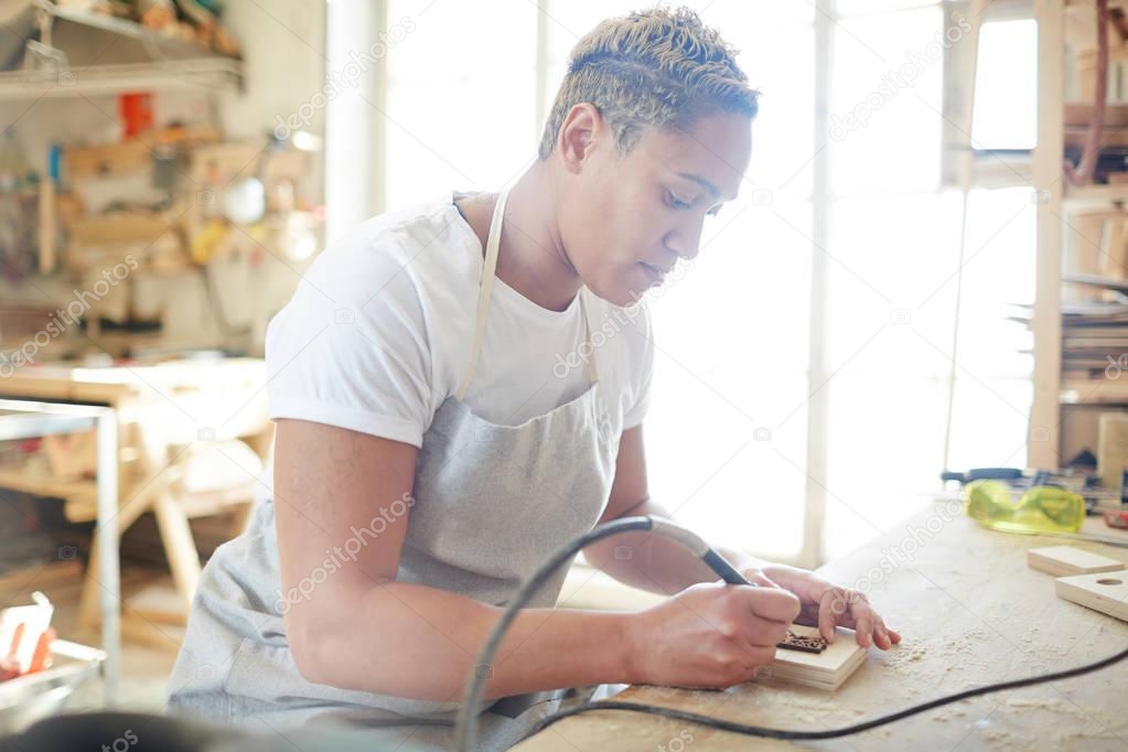 Young specialist with special pyrographic equipment making burnt ornament and carves on wooden plank