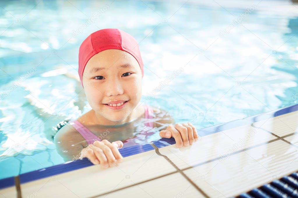 Cute little girl in swim-cap looking at camera during training in swimming-pool