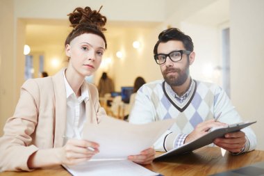 Genç ciddi adam ve kadın toplantı sırasında kameraya bakarak belgelerle