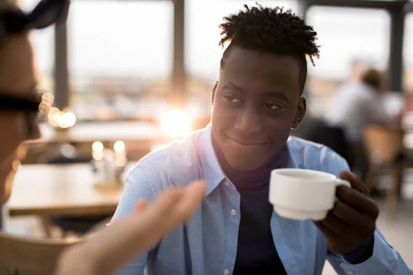 Friendly young African man with cup of coffee listening to explanation of colleague in cafe