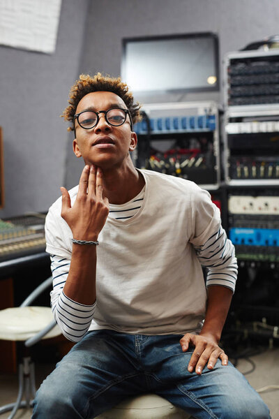 Young cool guy in casualwear and eyeglasses making gesture with his fingertips behind chin