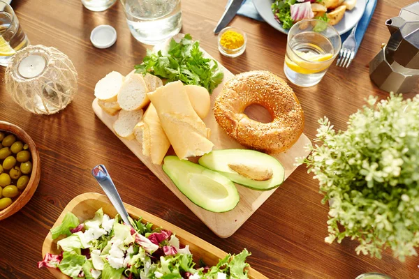 新鲜的面包和鳄梨一半在木板周围的沙拉 柠檬水 蜡烛和其他食物桌上 — 图库照片