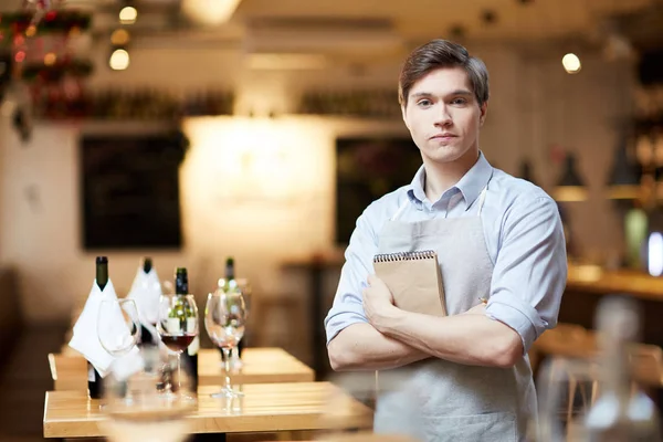 Young waiter with notepad looking at camera by one of tables with wine