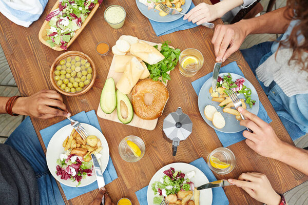 Обзор здорового домашнего завтрака на деревянном столе и человеческих рук с вилками и ножами над тарелками с едой
