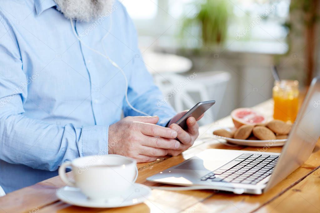 Mobile senior man with smartphone texting by table in cafe with laptop in front