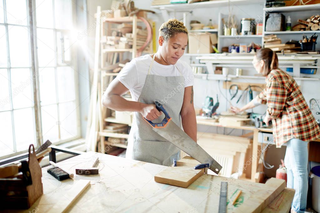 Young woman in apron sawing wood with her colleague working on background