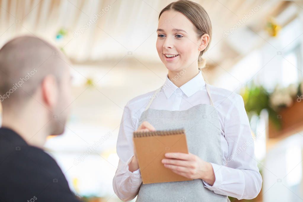 Young friendly waitress with notepad listening to her client order in restaurant in cafe