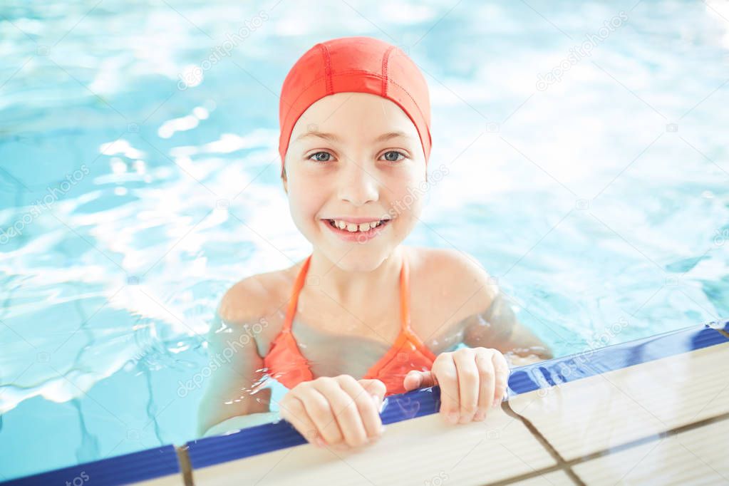 Smiling schoolgirl in swim-cap looking out of water while training in swimming-pool