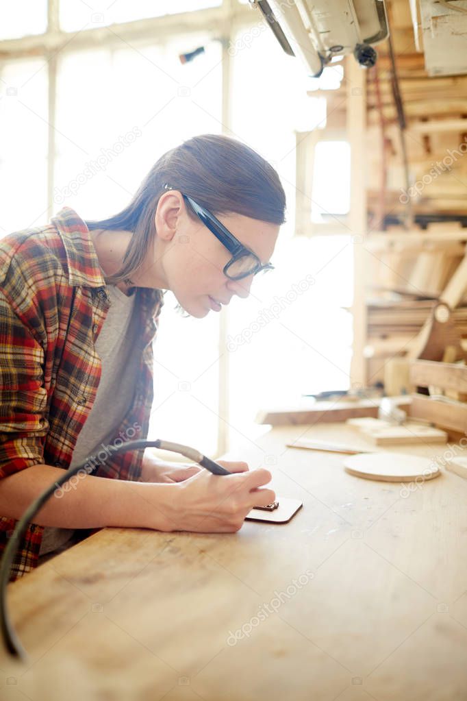Young female carpenter with pyrographic equipment drawing ornaments on wooden board